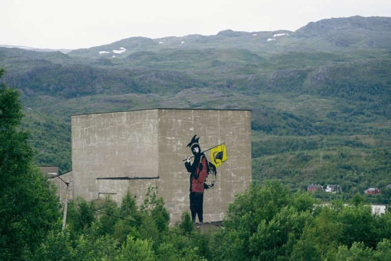 Anorakk, ryggsekk, samisk lue og Nordlands fylkesvåpen på en smuldrende betongvegg i Sulitjelma, del av det første smelteverket nedlagt i 1912. Graffitien skal illustrere Mons Petter. © Marie Nystad Helgesen